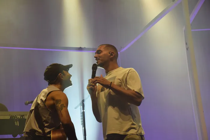 Ein Sänger im weißen T-Shirt singt in ein Mikrofon, während ein Musiker mit Kappe und Rosen-Tattoo ihm gegenübersteht.