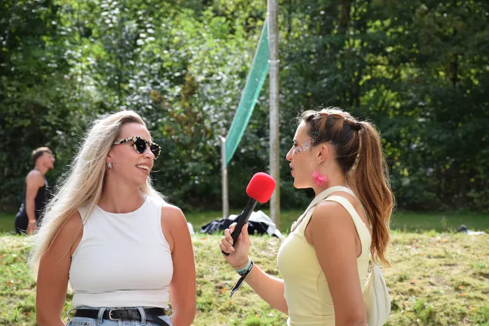 Eine Frau mit Glitzer im Gesicht hält ein rotes Mikrofon zu einer lächelnden Frau mit Sonnenbrille und weißem Top.