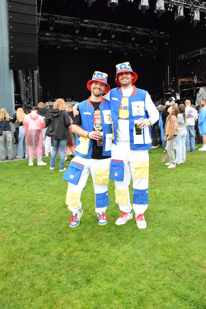 Zwei lächelnde Männer in blau-gelben Outfits und roten Hüten halten Getränke vor einer Bühne.