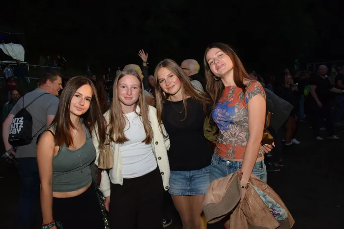 Vier junge Frauen lächeln in die Kamera und posieren in einer Menschenmenge bei einer nächtlichen Veranstaltung.