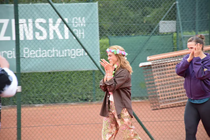 Eine Frau mit Blumenkranz klatscht auf einem Tennisplatz bei der Jubiläumsfeier der MTC/TC Halver Open.
