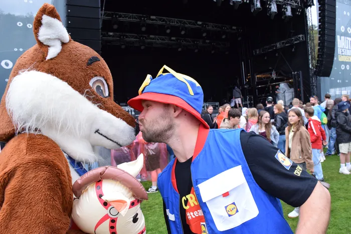 Ein Mann in einer blauen Crew-Weste und einem rot-blauen Hut lehnt sich an die Nase eines großen Fuchs-Maskottchens vor einer Festivalbühne.