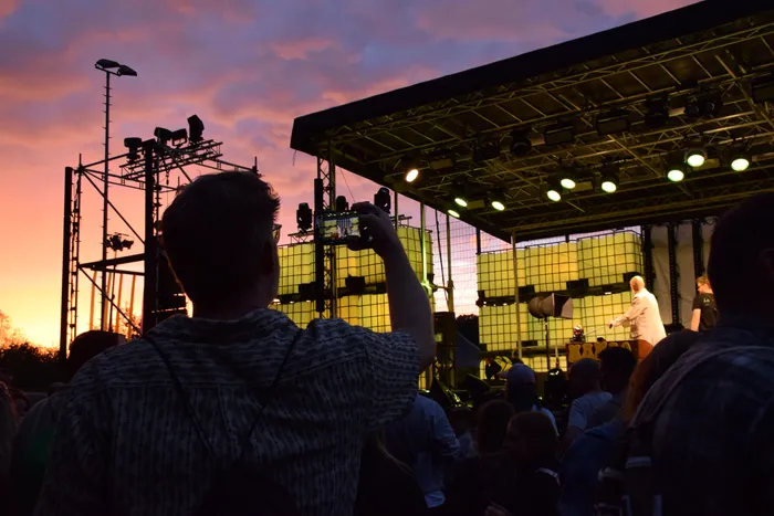 Eine Person fotografiert eine Konzertbühne mit leuchtend gelben Paneelen und einem Musiker bei Sonnenuntergang.
