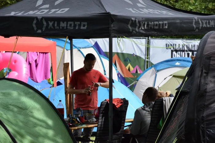 Ein Mann in einem roten T-Shirt bereitet unter einem schwarzen XLMOTO-Pavillon etwas vor, im Hintergrund stehen bunte Campingzelte.
