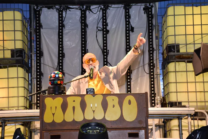 Ein Mann mit orangefarbener Sonnenbrille singt hinter einem Pult mit der Aufschrift „MAMBO“ in ein Mikrofon.