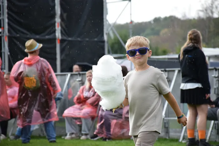 Ein Junge mit blauen Sonnenbrillen hält eine große weiße Zuckerwatte, während im Hintergrund Menschen in Regenjacken stehen.