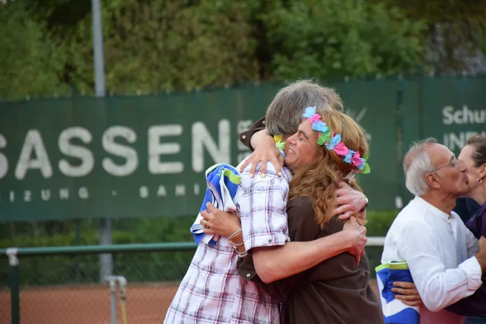 Eine Frau mit einem bunten Blumenkranz umarmt einen Mann bei einer Jubiläumsfeier auf einer Tennisanlage.