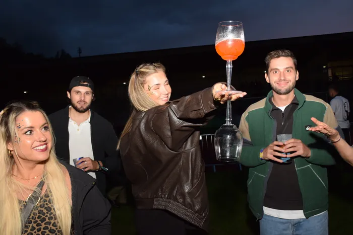 Eine junge Frau in einer braunen Lederjacke hält ein großes Glas mit einem orangefarbenen Getränk hoch, während sie mit anderen Menschen bei einer Abendveranstaltung feiert.
