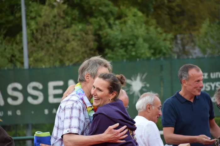 Eine Frau in einem lila Oberteil wird bei der Jubiläumsfeier der MTC/TC Halver Open von einer Person mit bunter Blumenkette umarmt.