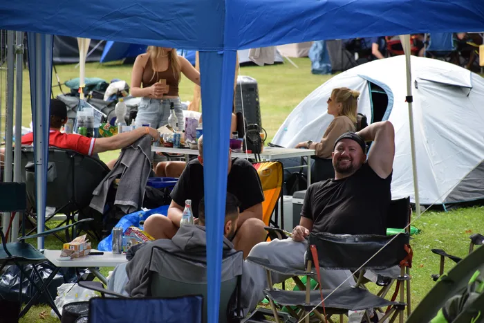 Menschen entspannen unter einem blauen Zeltdach auf einer Wiese vor einem weißen Zelt; ein lächelnder Mann sitzt in einem Campingstuhl.