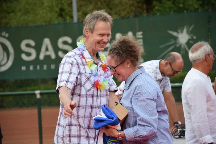 Ein lachender Mann mit einer bunten Blumenkette und eine Frau in einem blau gestreiften Hemd bei einer Jubiläumsfeier auf einer Tennisanlage.