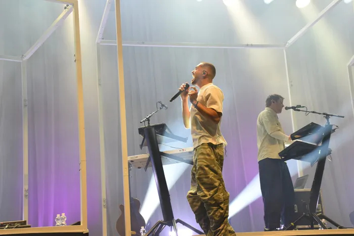 Ein Sänger der Band ClockClock singt in ein Mikrofon auf einer Bühne, während im Hintergrund ein Musiker am Keyboard spielt.