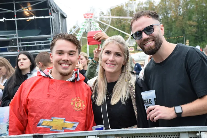 Drei lächelnde junge Menschen posieren bei einem Festival vor einem Riesenrad; einer trägt ein rotes Manchester United Sweatshirt.