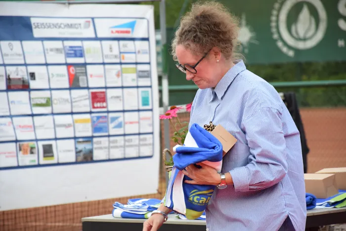 Eine Frau mit Brille und blau gestreiftem Hemd hält ein blaues Tuch und ein Paket vor einem Banner mit Sponsorenlogos.