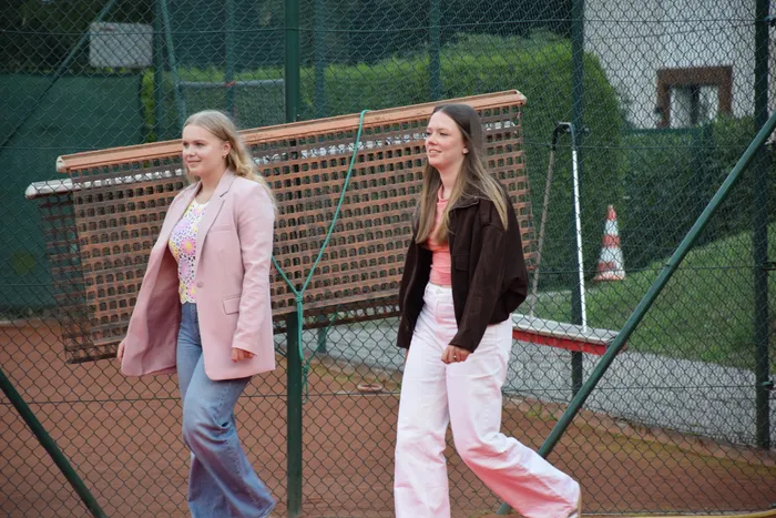 Zwei junge Frauen gehen über einen Tennisplatz vor einem Maschendrahtzaun; eine trägt einen rosa Blazer, die andere eine dunkle Jacke.
