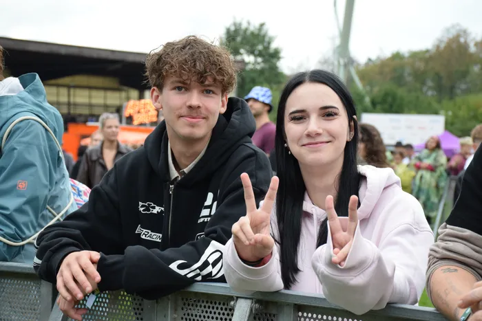 Ein junger Mann und eine junge Frau lächeln und zeigen Peace-Zeichen, während sie an einer Metallabsperrung beim Bautz-Samstag lehnen.