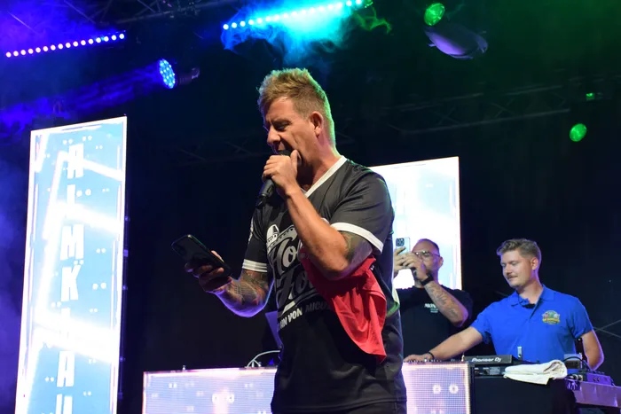 Almklausi singt in ein Mikrofon und hält ein Smartphone in der Hand auf einer Bühne mit farbiger Beleuchtung.