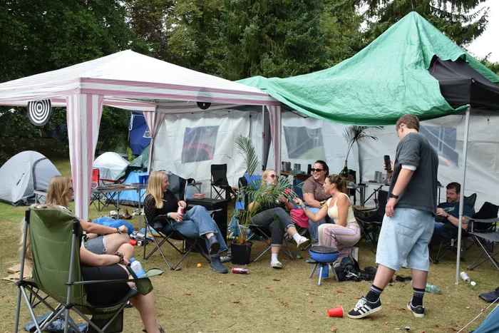 Eine Gruppe von Menschen sitzt in Campingstühlen unter einem Pavillon und einer grünen Plane auf einer Wiese vor Zelten.