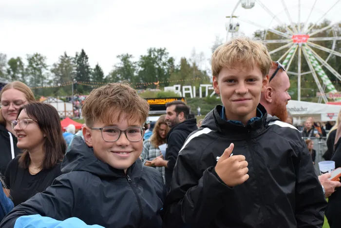 Zwei lächelnde Jungen, einer davon mit erhobenem Daumen, vor einem Riesenrad auf einem Volksfest.