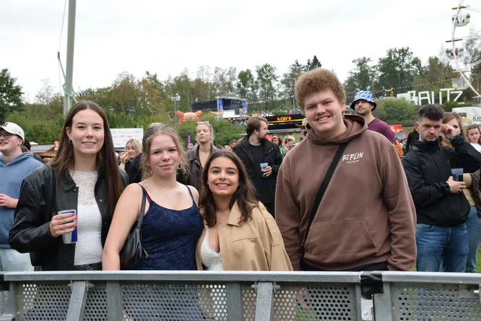 Eine Gruppe junger Menschen lächelt hinter einer Metallabsperrung bei einer Open-Air-Veranstaltung.
