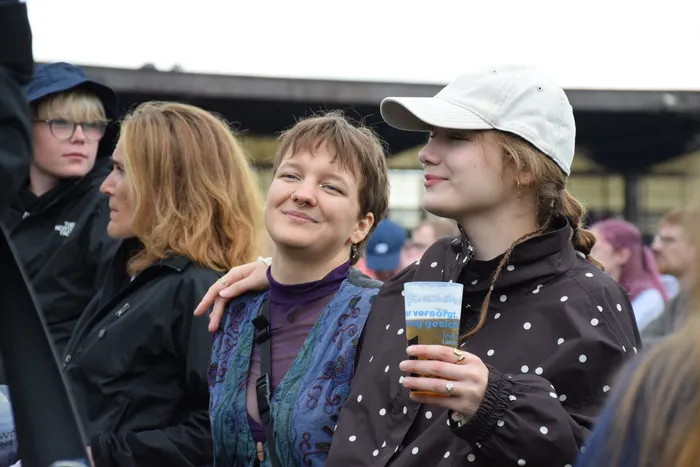 Zwei lächelnde Frauen in Regenkleidung stehen bei einer Veranstaltung im Freien, eine trägt eine weiße Kappe und hält einen Becher.