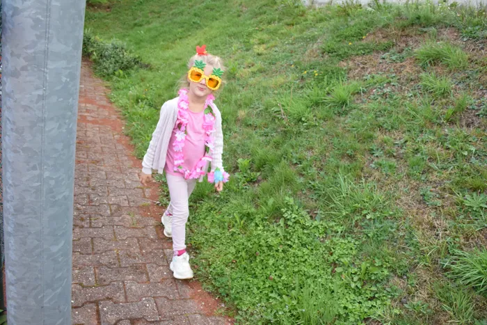 Ein kleines Mädchen mit gelben Ananas-Sonnenbrillen und einer rosa Girlande läuft auf einem gepflasterten Weg neben einer Rasenfläche.
