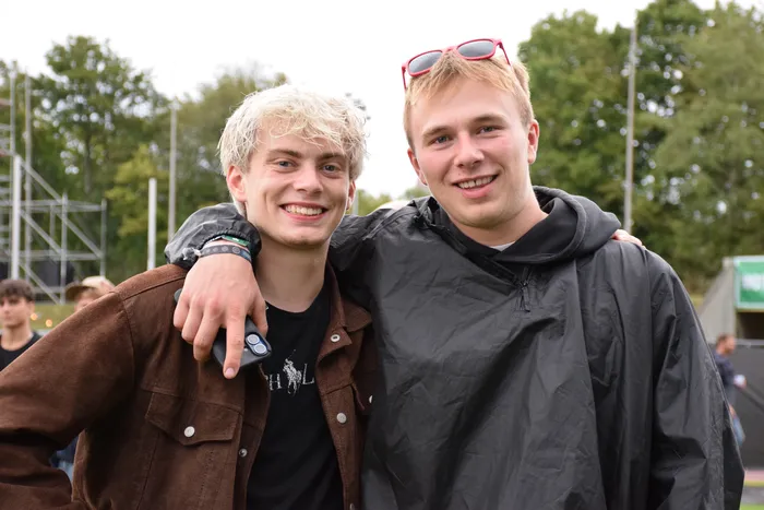 Zwei lächelnde junge Männer posieren eng umschlungen, einer trägt eine braune Jacke, der andere einen schwarzen Regenmantel mit roter Sonnenbrille.