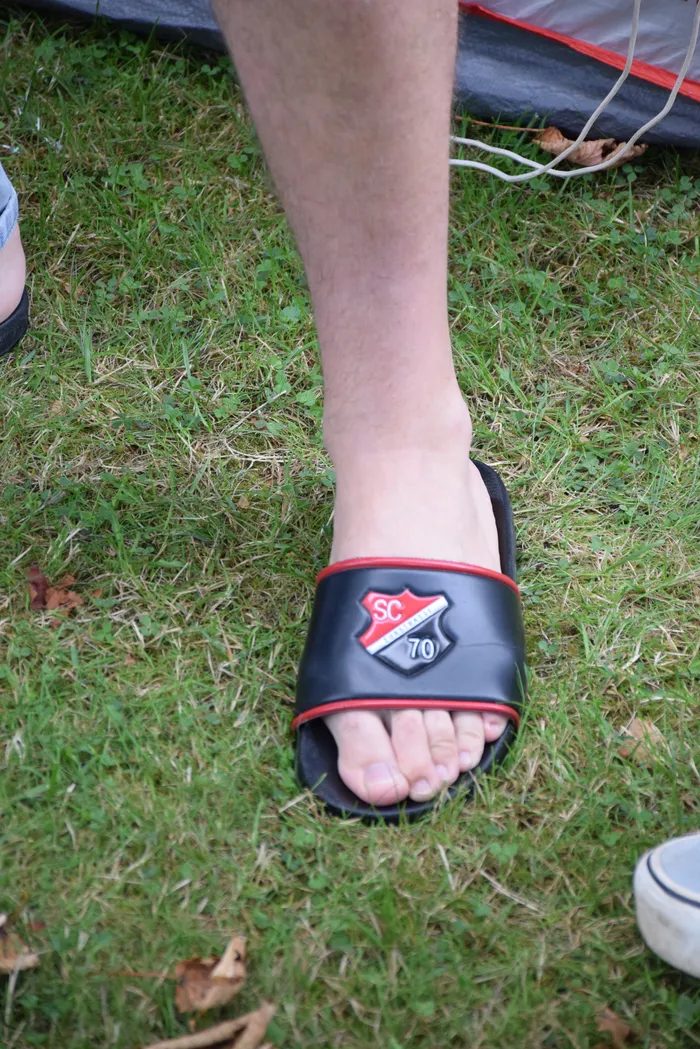 Ein Fuß in einem schwarzen Slipper mit rotem Rand und einem „SC 70“-Wappen auf einer Rasenfläche.