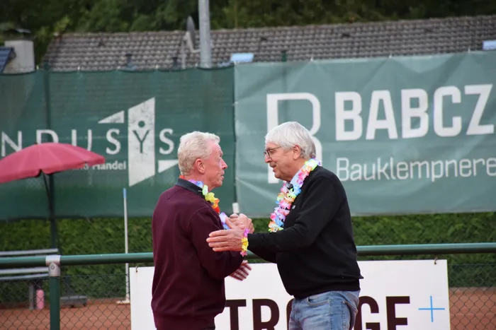 Zwei Männer mit bunten Blumenketten schütteln sich die Hände auf einem Tennisplatz während einer Jubiläumsfeier.