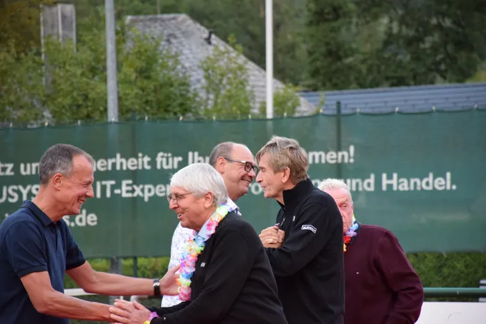 Menschen lachen und begrüßen sich bei der Jubiläumsfeier der MTC/TC Halver Open. Eine Frau trägt eine bunte Blumenkette.