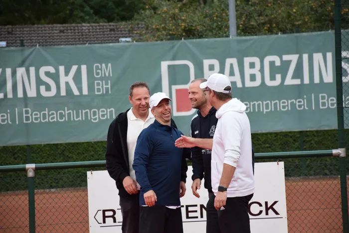 Vier Männer unterhalten sich lachend auf einem Tennisplatz vor einem grünen Werbebanner.