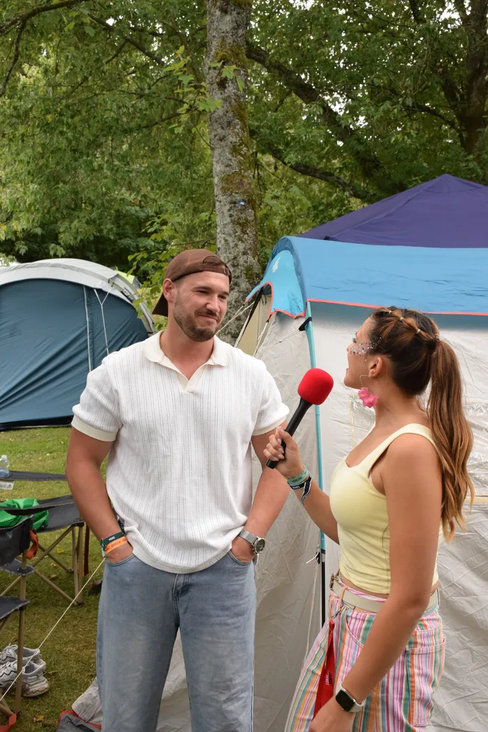 Eine Frau mit Glitzer im Gesicht interviewt einen Mann in einem weißen Poloshirt und brauner Kappe mit einem roten Mikrofon vor Zelten.