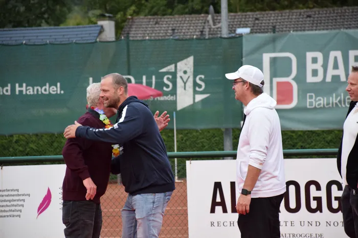 Zwei Männer umarmen sich auf einem Tennisplatz, einer trägt eine bunte Blumenkette um den Hals.
