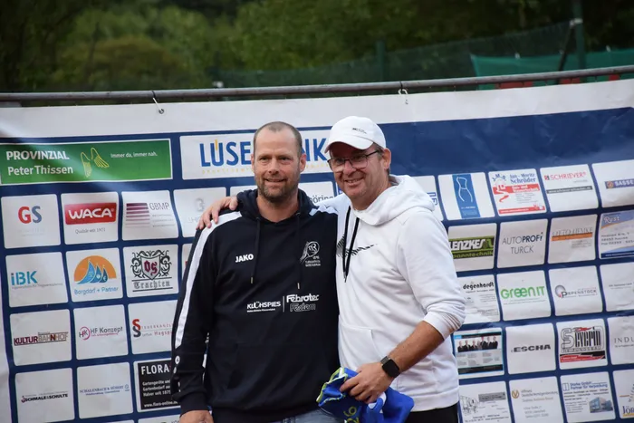 Zwei Männer lächeln vor einer Sponsorenwand bei der Feier zum 20-jährigen Jubiläum der MTC/TC Halver Open.
