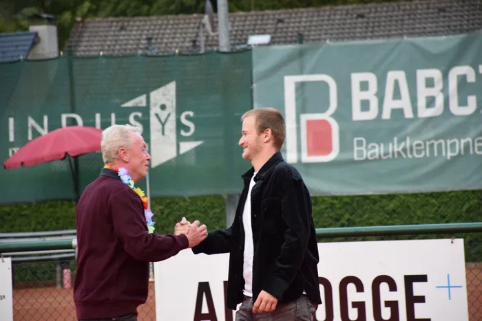 Zwei Männer schütteln sich auf einer Tennisanlage die Hände, einer trägt eine bunte Blumenkette.