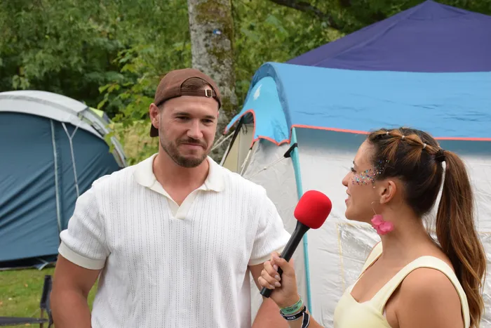 Eine Frau mit Glitzer im Gesicht interviewt einen Mann in weißem Poloshirt und brauner Kappe vor Zelten.