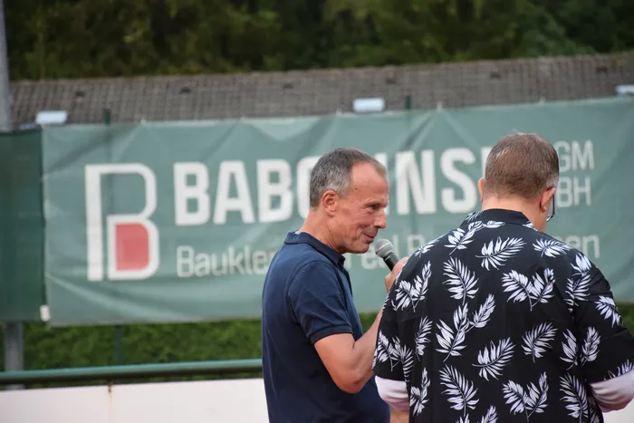 Ein Mann in einem dunkelblauen Poloshirt spricht in ein Mikrofon zu einem Mann in einem schwarz-weiß gemusterten Hemd vor einem grünen Banner.