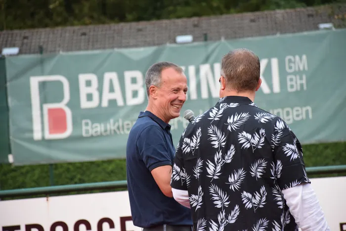 Ein lächelnder Mann in einem blauen Poloshirt wird von einer Person in einem schwarz-weiß gemusterten Hemd mit einem Mikrofon interviewt.
