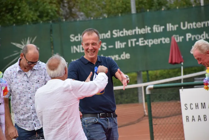 Ein lachender Mann im dunkelblauen Poloshirt begrüßt einen Mann im weißen Hemd auf einem Tennisplatz während der Jubiläumsfeier der MTC/TC Halver Open.