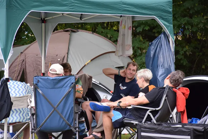 Eine Gruppe von Menschen sitzt in Campingstühlen unter einem grünen Pavillon vor einem großen Zelt.