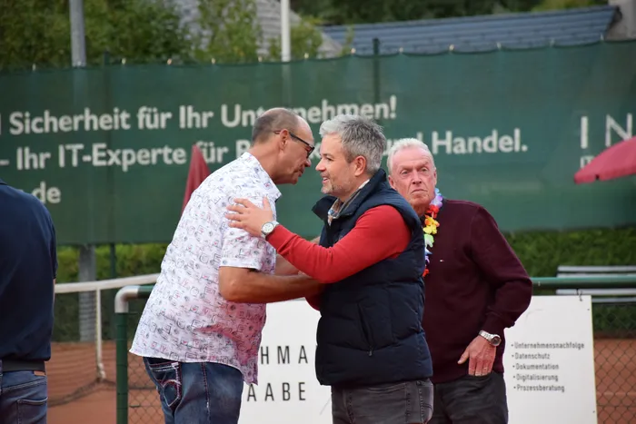 Zwei Männer im Gespräch auf einem Tennisplatz, ein dritter Mann mit einer bunten Blumenkette steht daneben.