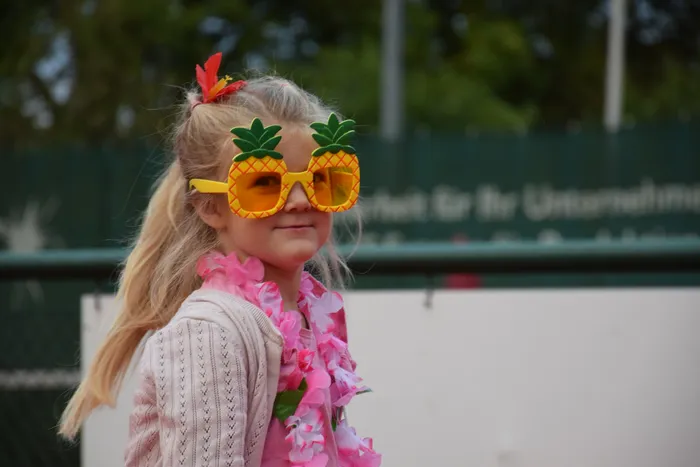Ein junges Mädchen mit gelber Ananas-Sonnenbrille und einer rosa Blumenkette lächelt.