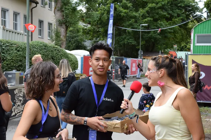 Eine Frau mit einem roten Mikrofon interviewt einen jungen Mann, während sie gemeinsam eine Pappschachtel halten.