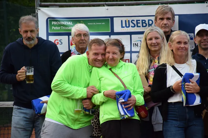 Eine Gruppe lächelnder Menschen, darunter ein Paar in leuchtend grünen Jacken, feiert das Jubiläum der MTC/TC Halver Open vor einem Sponsoren-Banner.
