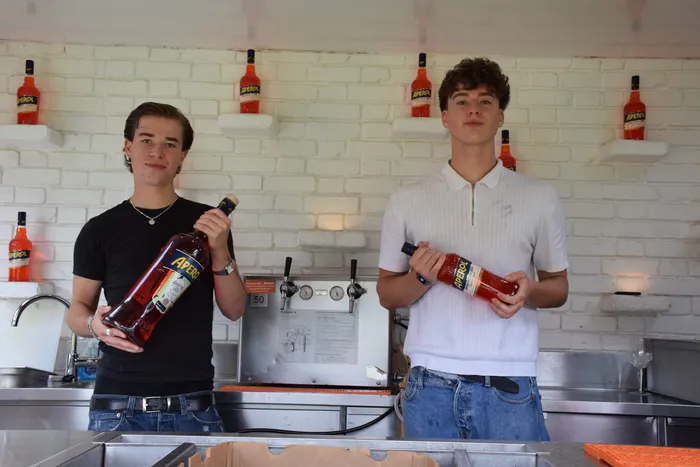 Zwei junge Männer halten jeweils eine Flasche Aperol hinter einer Bar vor einer weißen Ziegelwand mit weiteren Aperol-Flaschen.
