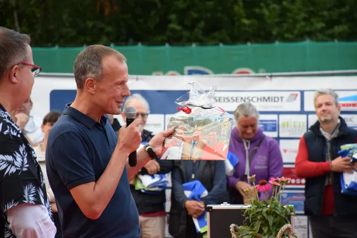 Ein Mann in einem blauen Poloshirt spricht in ein Mikrofon und hält ein verpacktes Geschenk während der Jubiläumsfeier der MTC/TC Halver Open hoch.