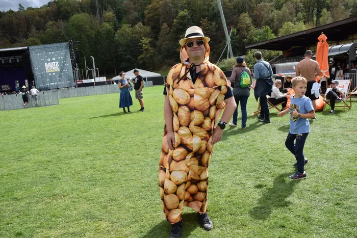 Ein Mann in einem Kartoffelsack-Kostüm mit Strohhut und Sonnenbrille steht auf einer Wiese bei einem Festival.
