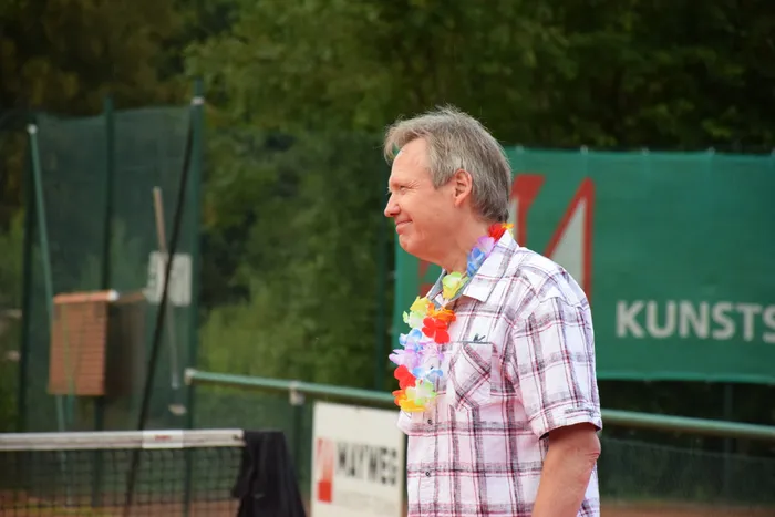 Ein lächelnder Mann in einem karierten Hemd trägt eine bunte Blumenkette auf einer Tennisanlage.