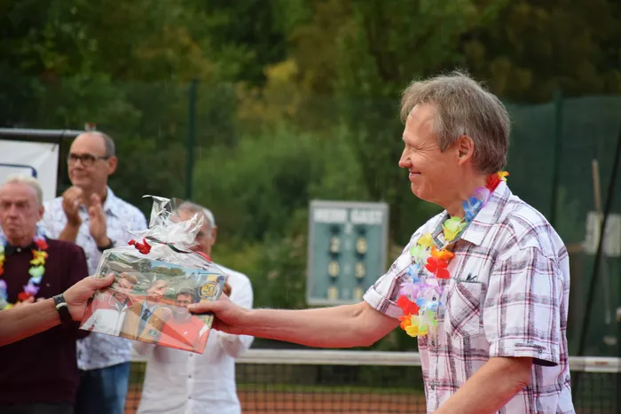 Ein Mann mit bunter Blumenkette erhält ein verpacktes Geschenk auf einer Tennisanlage während einer Jubiläumsfeier.