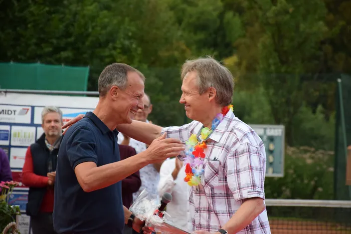 Zwei Männer lachen sich bei einer Jubiläumsfeier auf einer Tennisanlage zu, einer trägt eine bunte Blumenkette um den Hals.